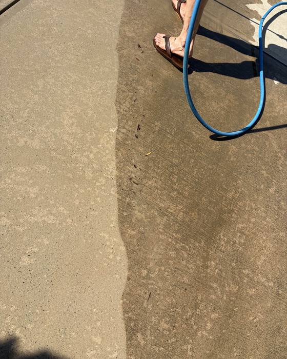 A patio slab mid-clean, with a garden hose coiled across it. A clear line runs down the middle between the washed and unwashed halves.